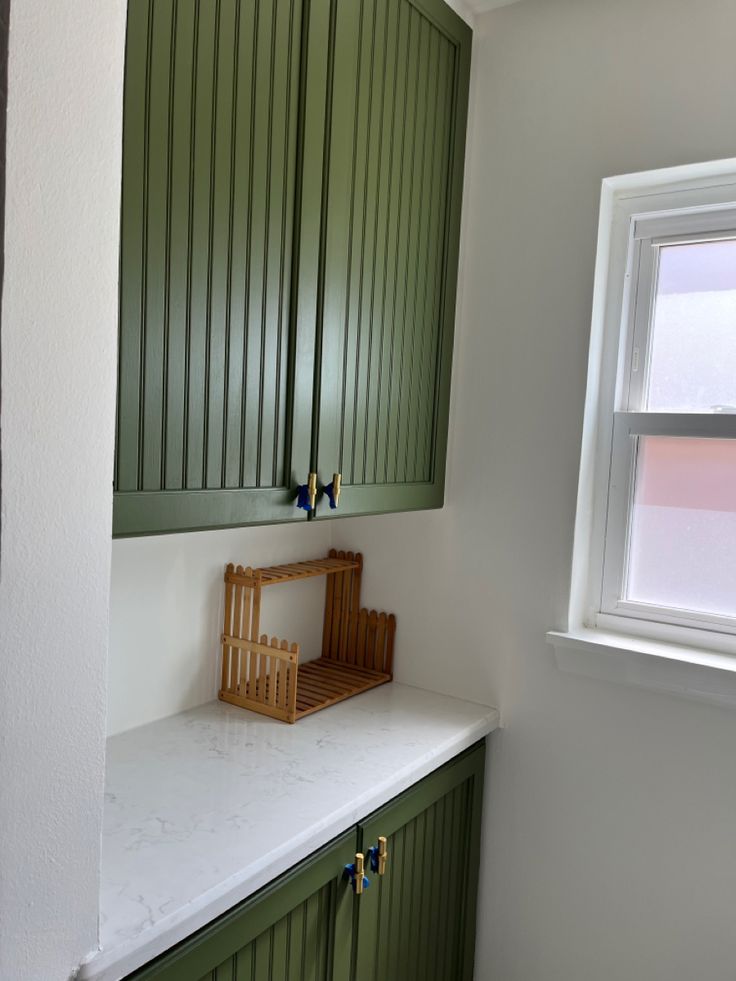 a kitchen with green cabinetry and white counter tops next to a window in the corner