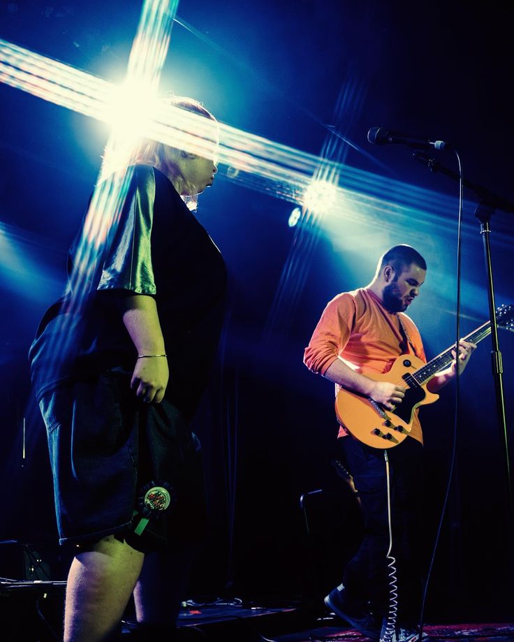 two people standing on stage with guitars and microphones in front of them, one playing the guitar