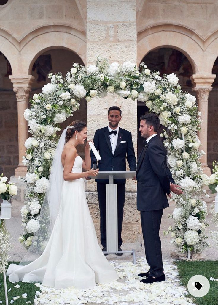 a bride and groom standing in front of an arch with white flowers on the grass