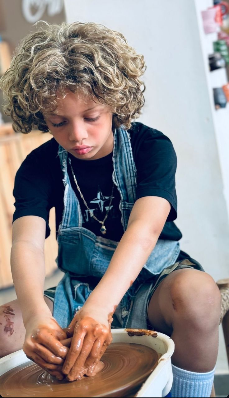 a little boy that is sitting in front of a pottery bowl with clay on it