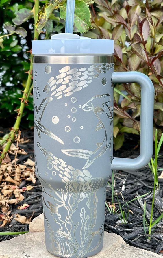 a stainless steel travel mug with an ocean scene on the lid and handle is sitting in front of some plants