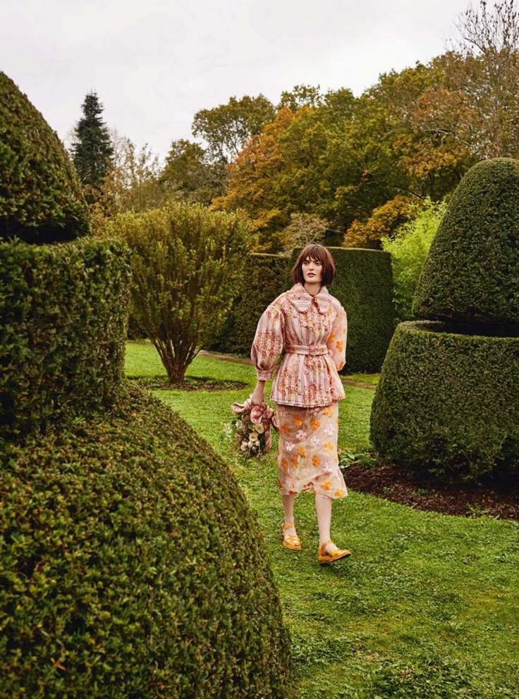 a woman is walking through the grass in front of some hedges and bushes with flowers