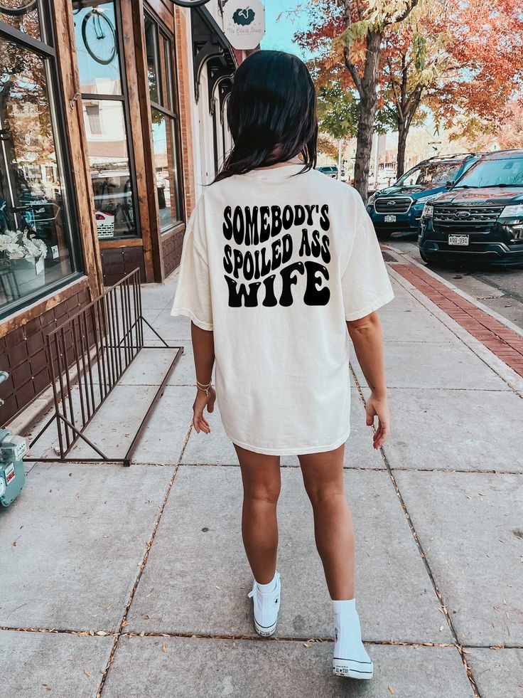 a woman walking down the sidewalk wearing an anti social wives club shirt on her back