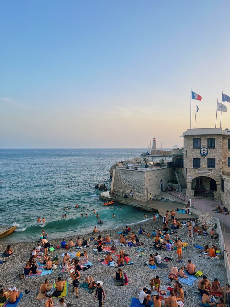 many people are sitting on the beach near the water and in front of an old building