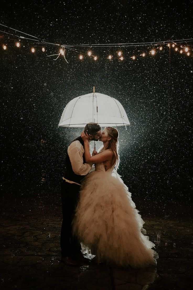 a bride and groom kissing under an umbrella in the rain with lights strung above them