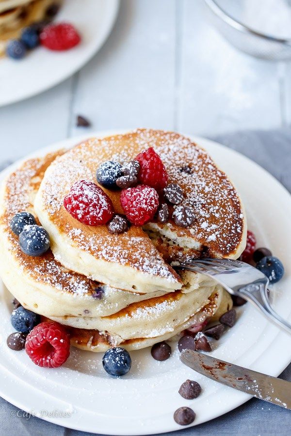 stack of pancakes with berries and powdered sugar on white plate next to silverware