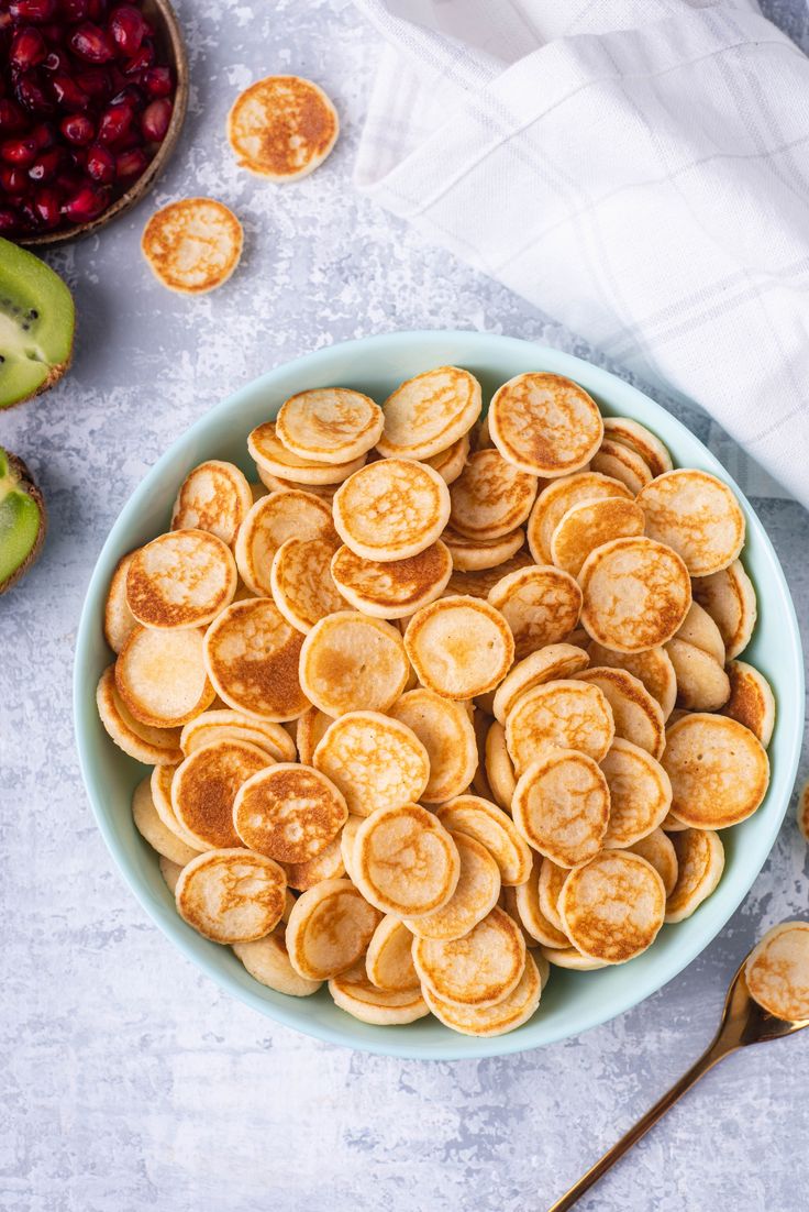 a blue bowl filled with pancakes next to apples and spoons on a white table