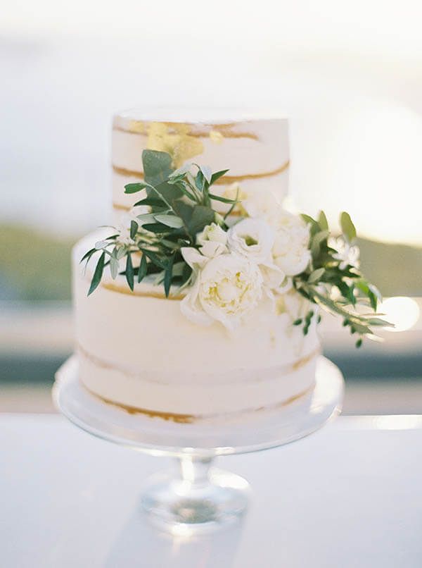 a wedding cake with white flowers and greenery sits on a clear pedestal in front of the ocean