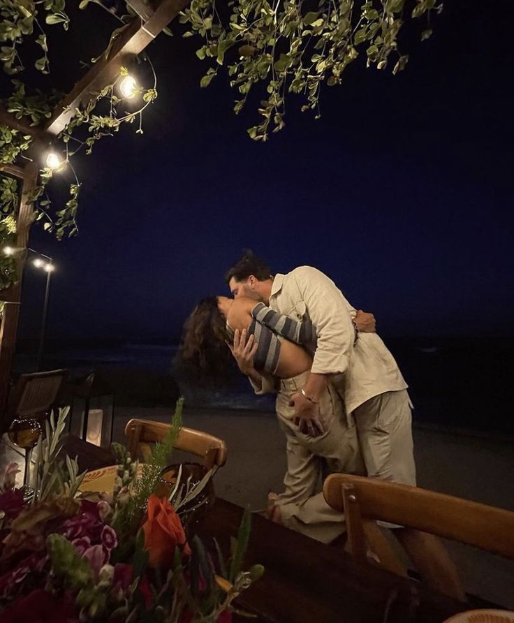 a man and woman kissing under an arbor with lights on the beach in the background