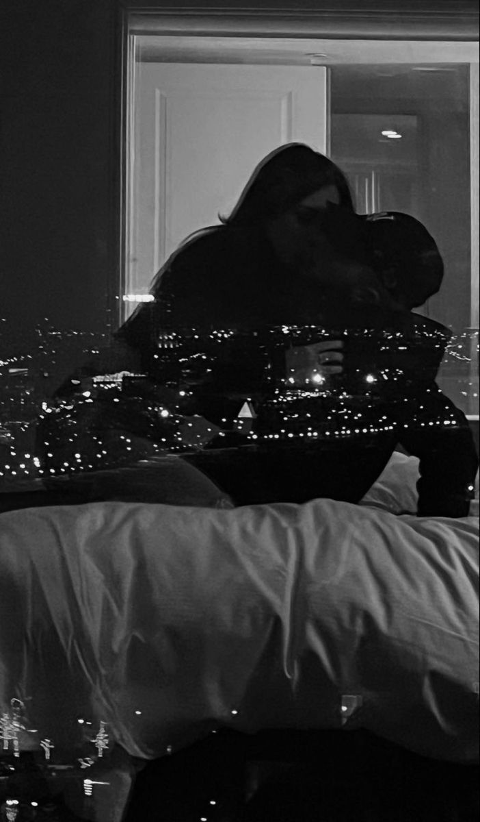 black and white photograph of woman kissing man in bed with city lights at night behind her