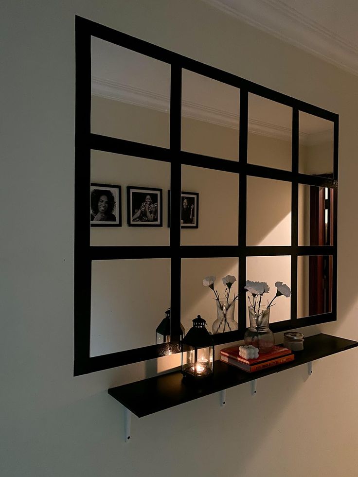 a mirror on the wall above a shelf with candles and flowers in it next to a window