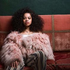 a woman sitting on top of a couch next to a red wall and wearing a pink fur coat