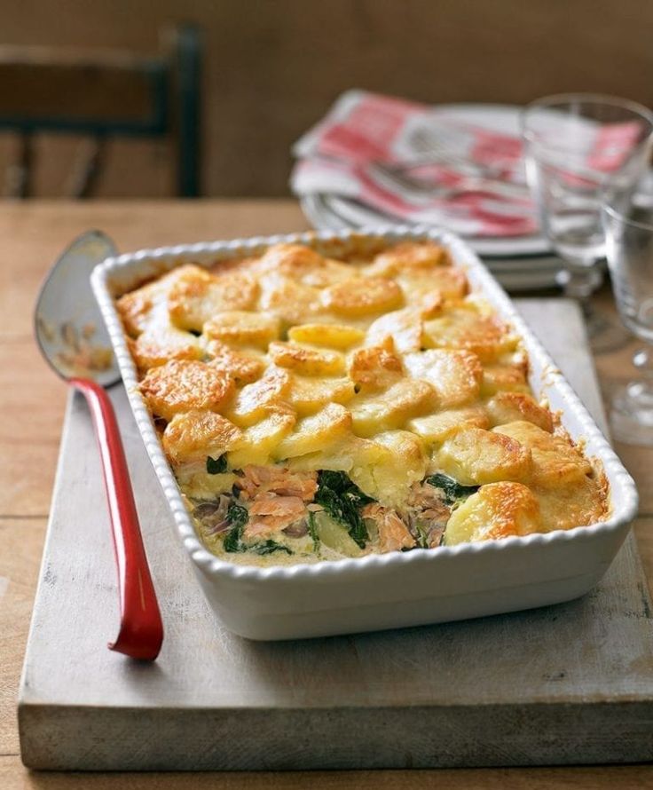 a casserole dish with potatoes and spinach on a wooden table next to utensils