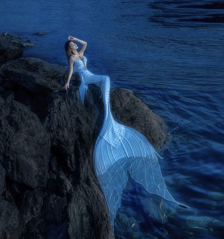 a woman in a blue dress is sitting on rocks by the water with her hands behind her head