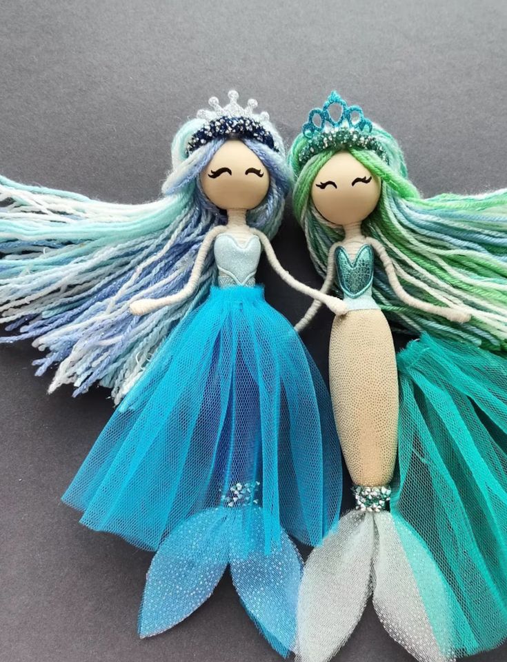 two little mermaid dolls sitting next to each other on top of a gray surface with blue and green hair