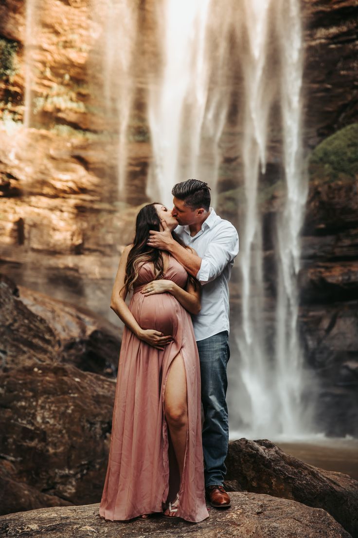 a pregnant couple kissing in front of a waterfall