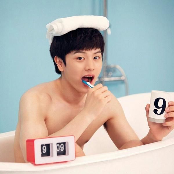 a young man brushing his teeth in the bathtub while holding an alarm clock and toothbrush