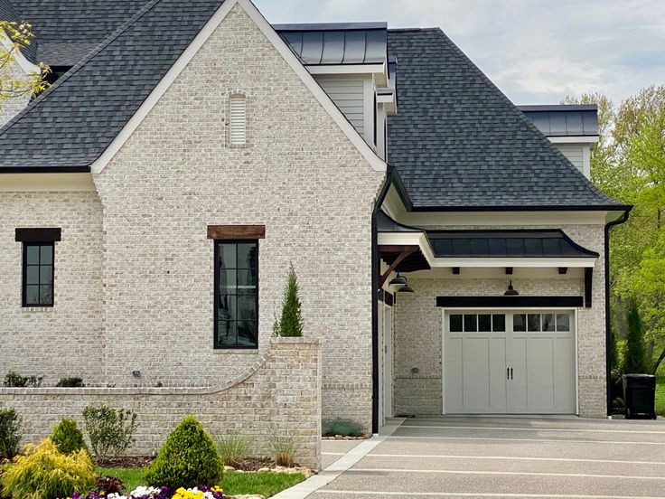 a white brick house with black shingles on the roof and two garages in front