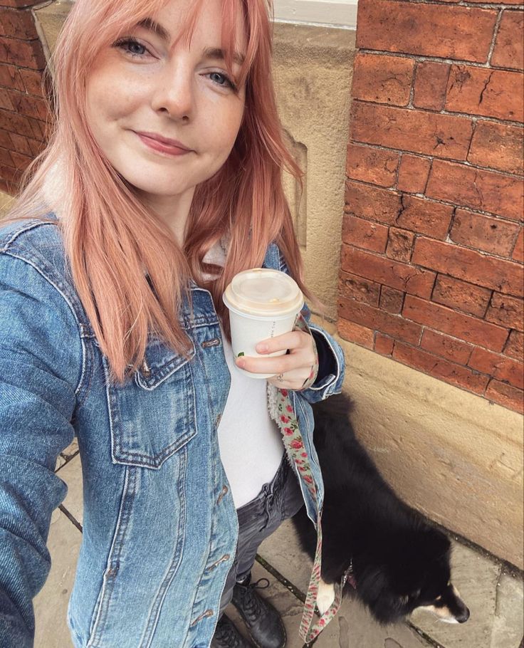 a woman with pink hair is holding a coffee cup in her hand and a black cat behind her