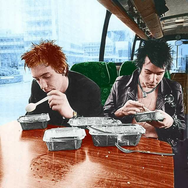 two young men sitting at a table eating from takeout containers with chopsticks