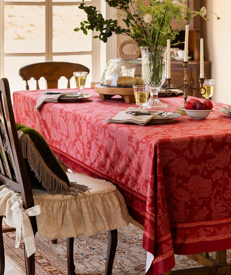 a red table cloth on a dining room table with two chairs and flowers in a vase