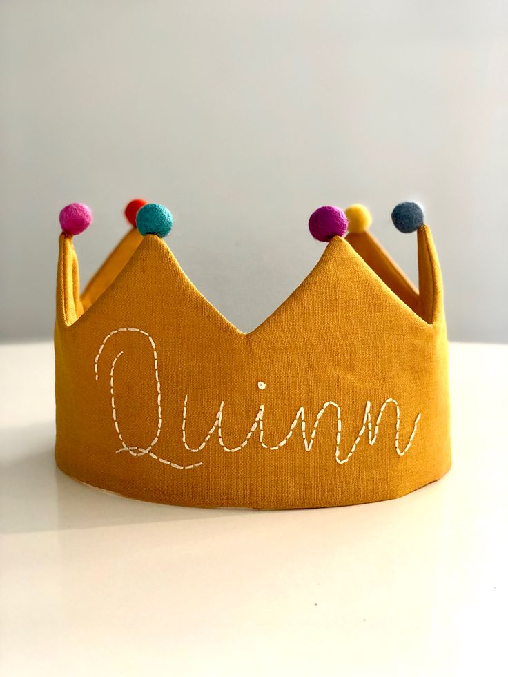 a yellow crown with the word queen written on it's side and pom - poms at the top