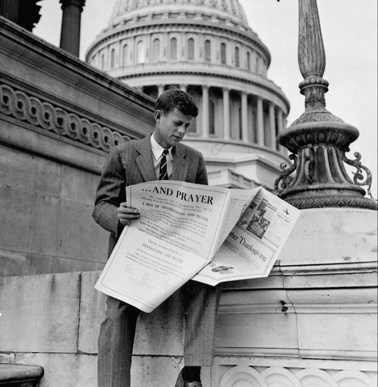 a man in a suit and tie standing on the steps reading a newspaper with the capital building in the background