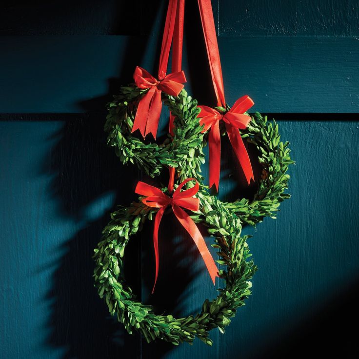 two wreaths with red bows hanging on a green door hang from a hook,