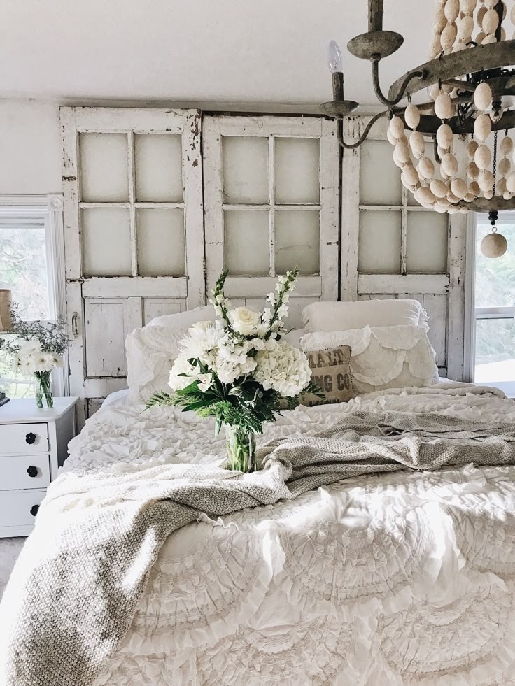 a white bed with flowers in a vase on the headboard and an old window behind it