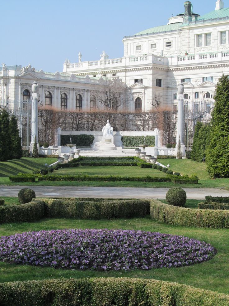 a large white building sitting next to a lush green park filled with purple and yellow flowers