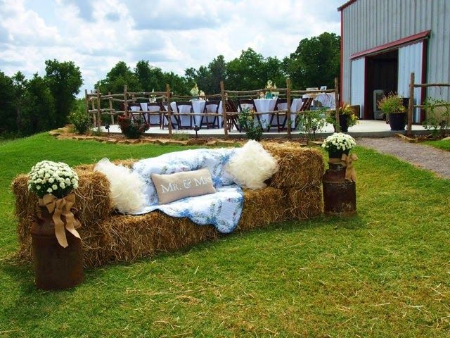 a hay couch with pillows and flowers on it in the middle of a field near a barn