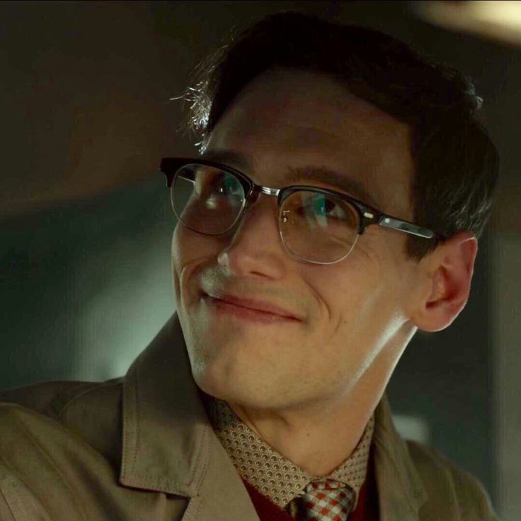 a man wearing glasses and a trench coat looks off to the side in a dimly lit room