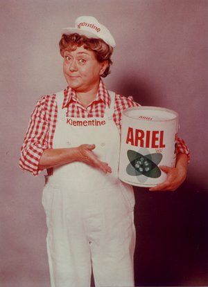 an older woman in white overalls holding a can of ariell with a green cat on it