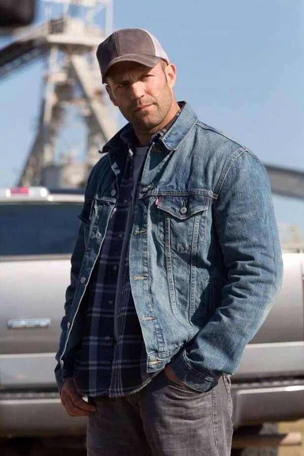 a man standing in front of a truck wearing a denim jacket and hat with his hands in his pockets