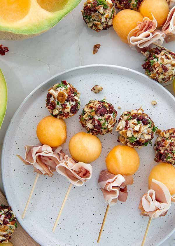 several appetizers are arranged on a white plate with fruit in the background, including melon and walnuts