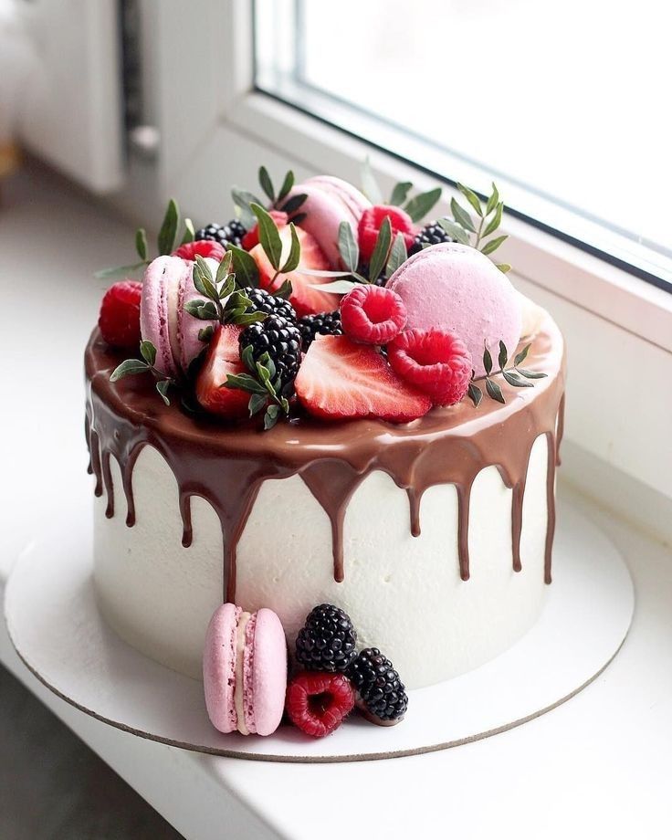 a white cake with chocolate icing, berries and macaroons on top sitting on a window sill