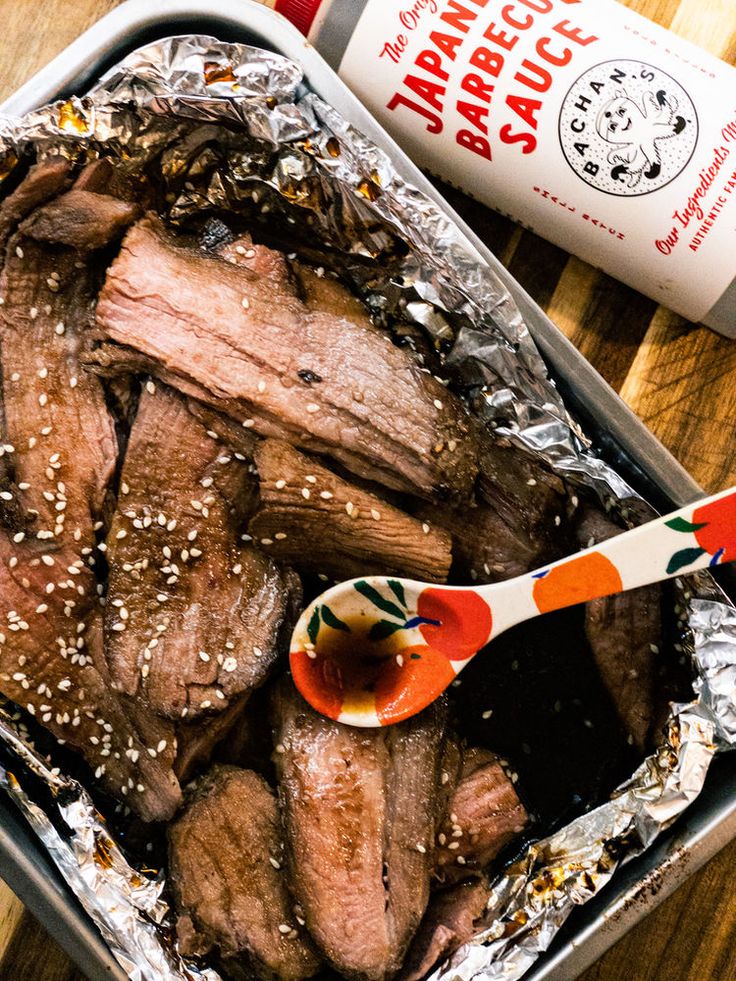 a tin with some meat in it next to a bottle of sauce and spoons