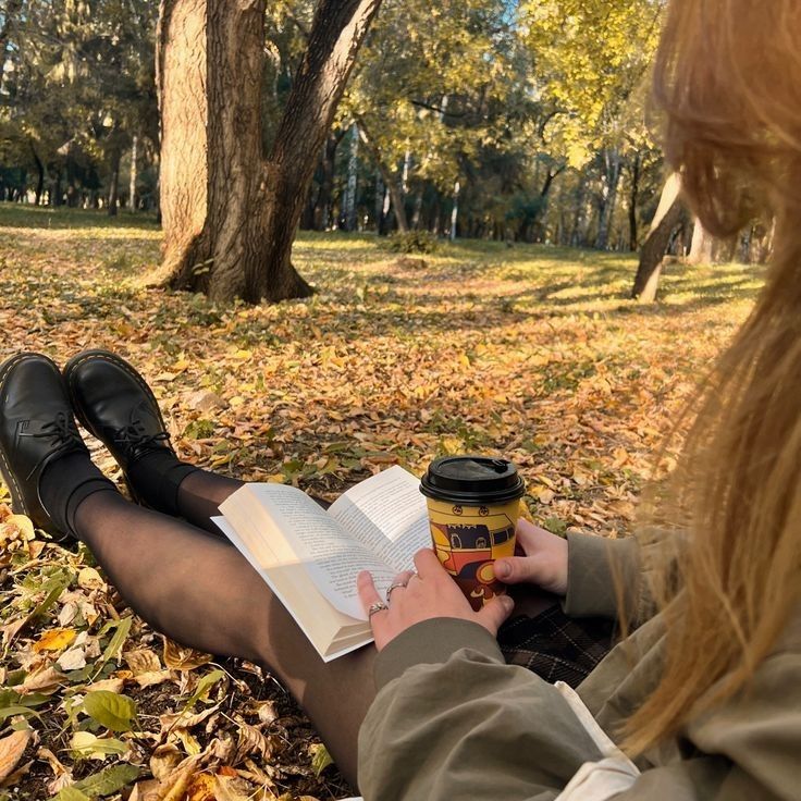 a woman sitting on the ground reading a book and holding a cup of coffee in her hand