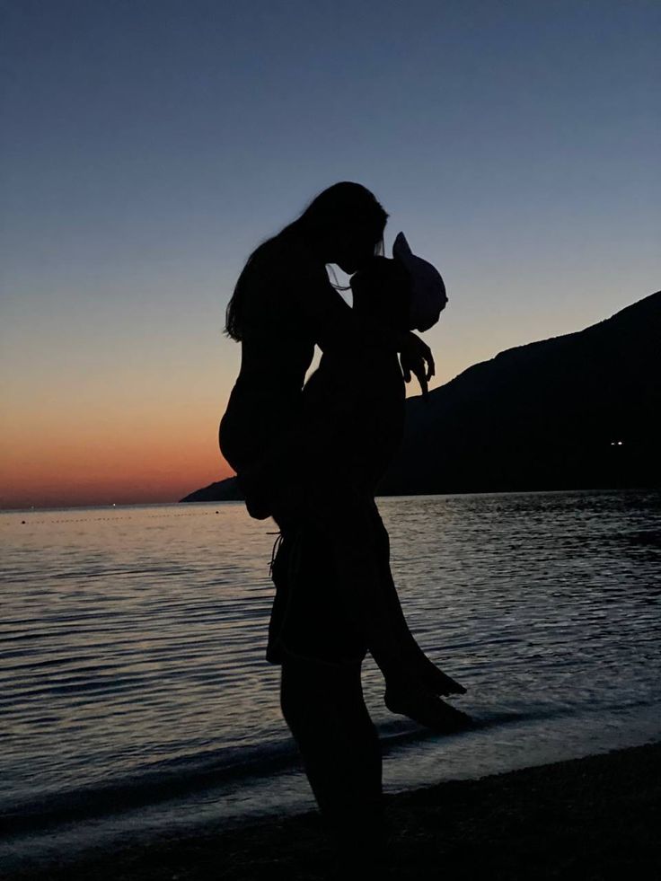 a woman standing on top of a beach next to the ocean holding a baby in her arms