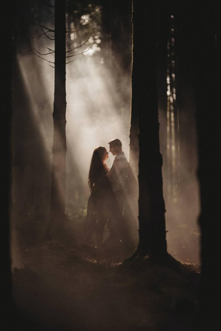 a man and woman standing in the woods with mist coming from behind them, surrounded by trees