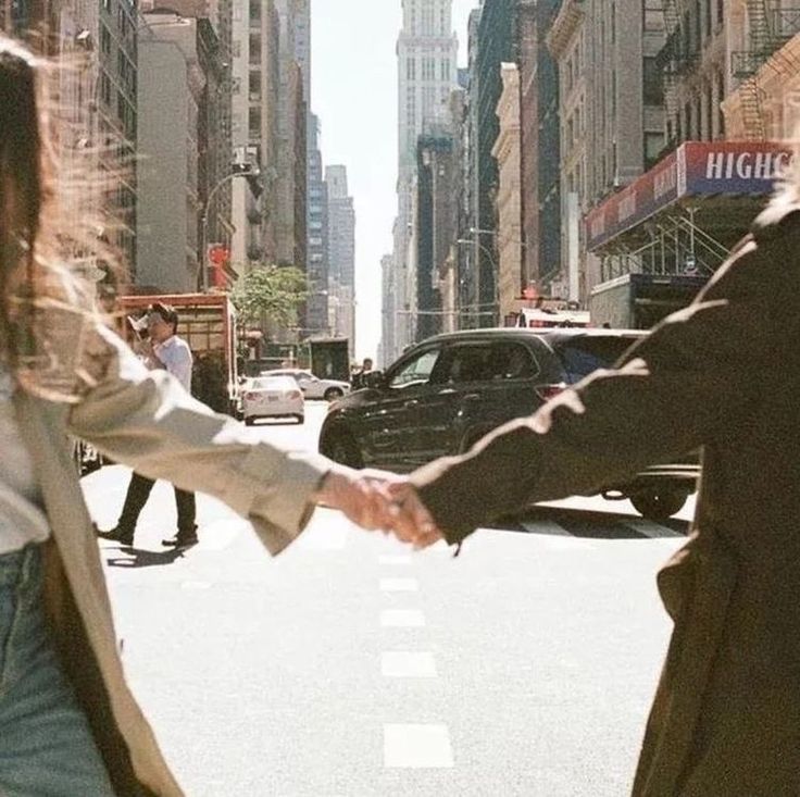 two people holding hands while walking down the street with cars and buildings in the background