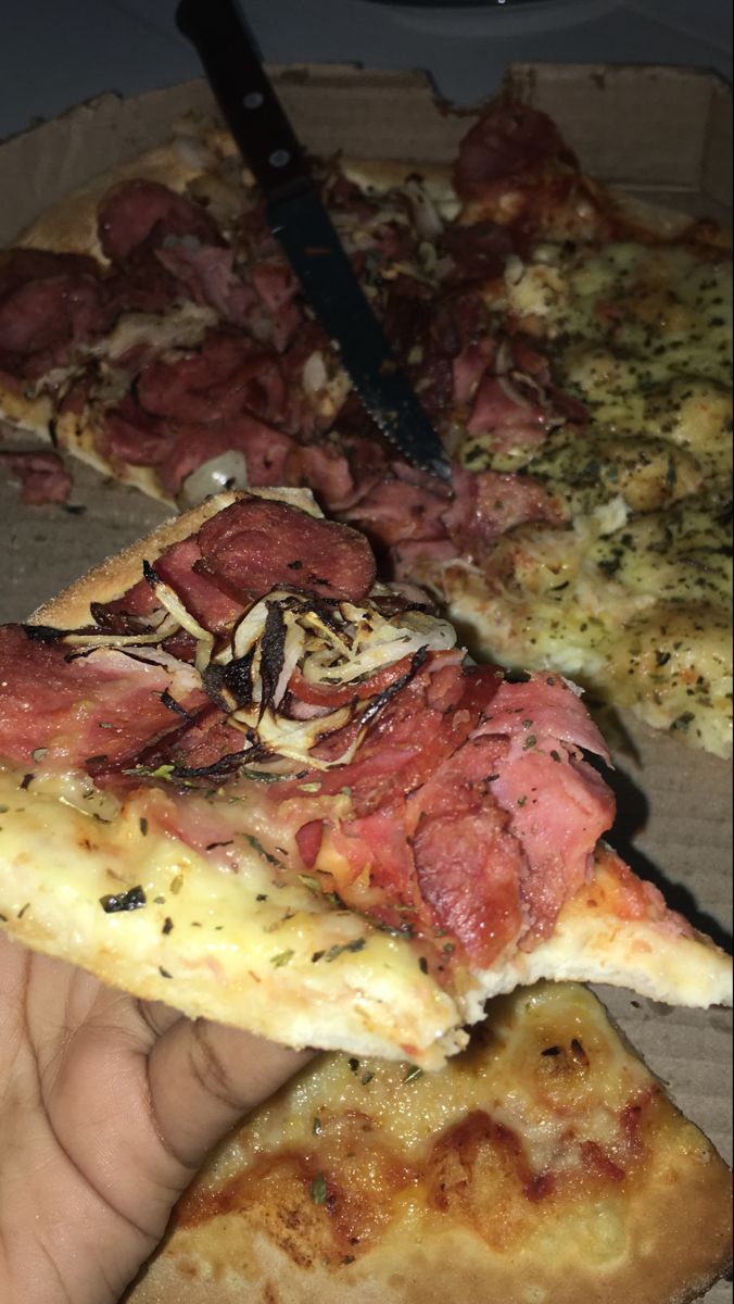 a person is holding two slices of pizza in front of the other half with meat on it