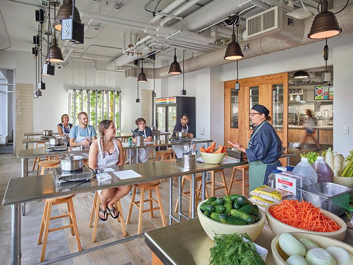 people are in the kitchen preparing food at tables with vegetables and fruits on them, while others watch