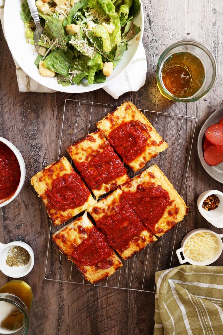 several square slices of pizza sitting on top of a wooden table next to bowls of salad