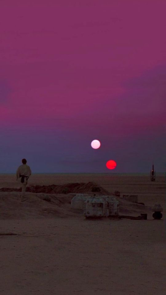 two people standing in the desert at dusk with red and pink clouds behind them,