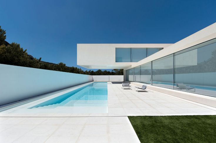 an empty swimming pool in front of a white building with glass walls and grass on the ground