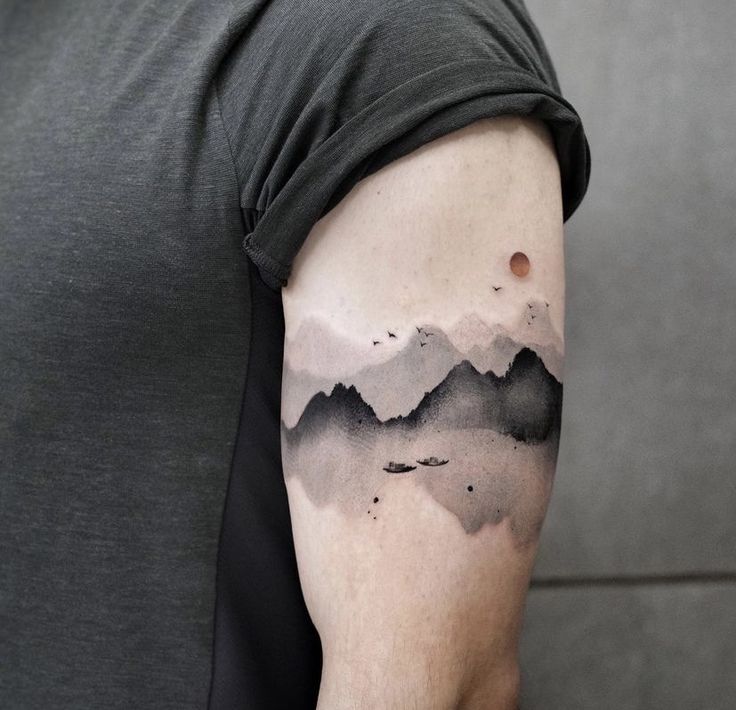 a man with a black and grey mountain tattoo on his left upper arm, in front of a gray wall