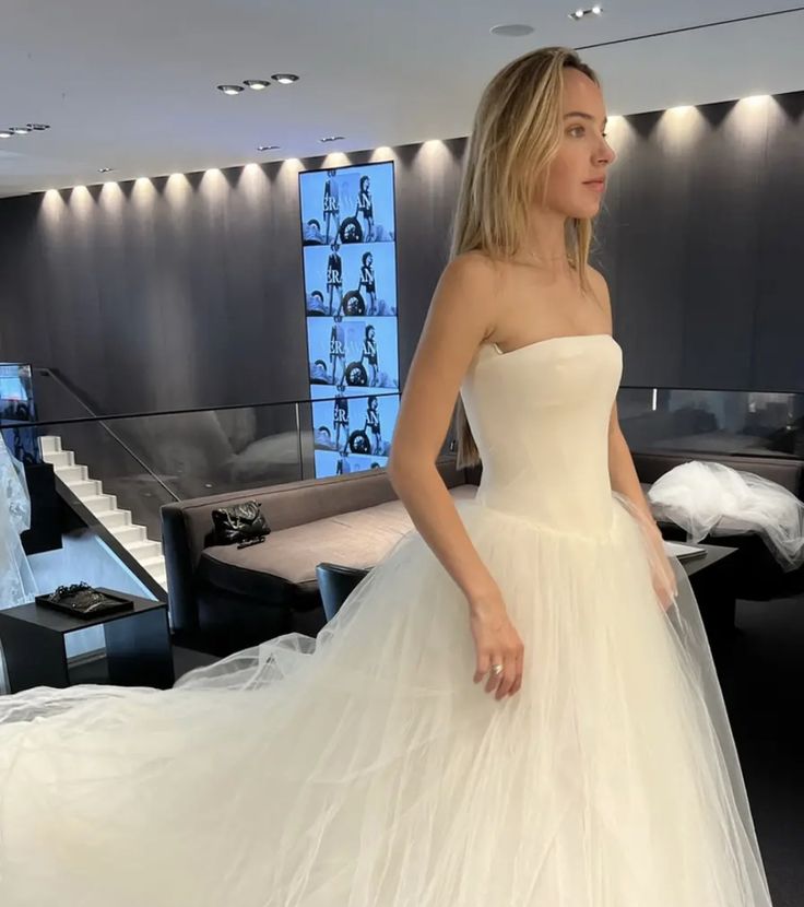 a woman in a white wedding dress standing next to a bed and looking off into the distance