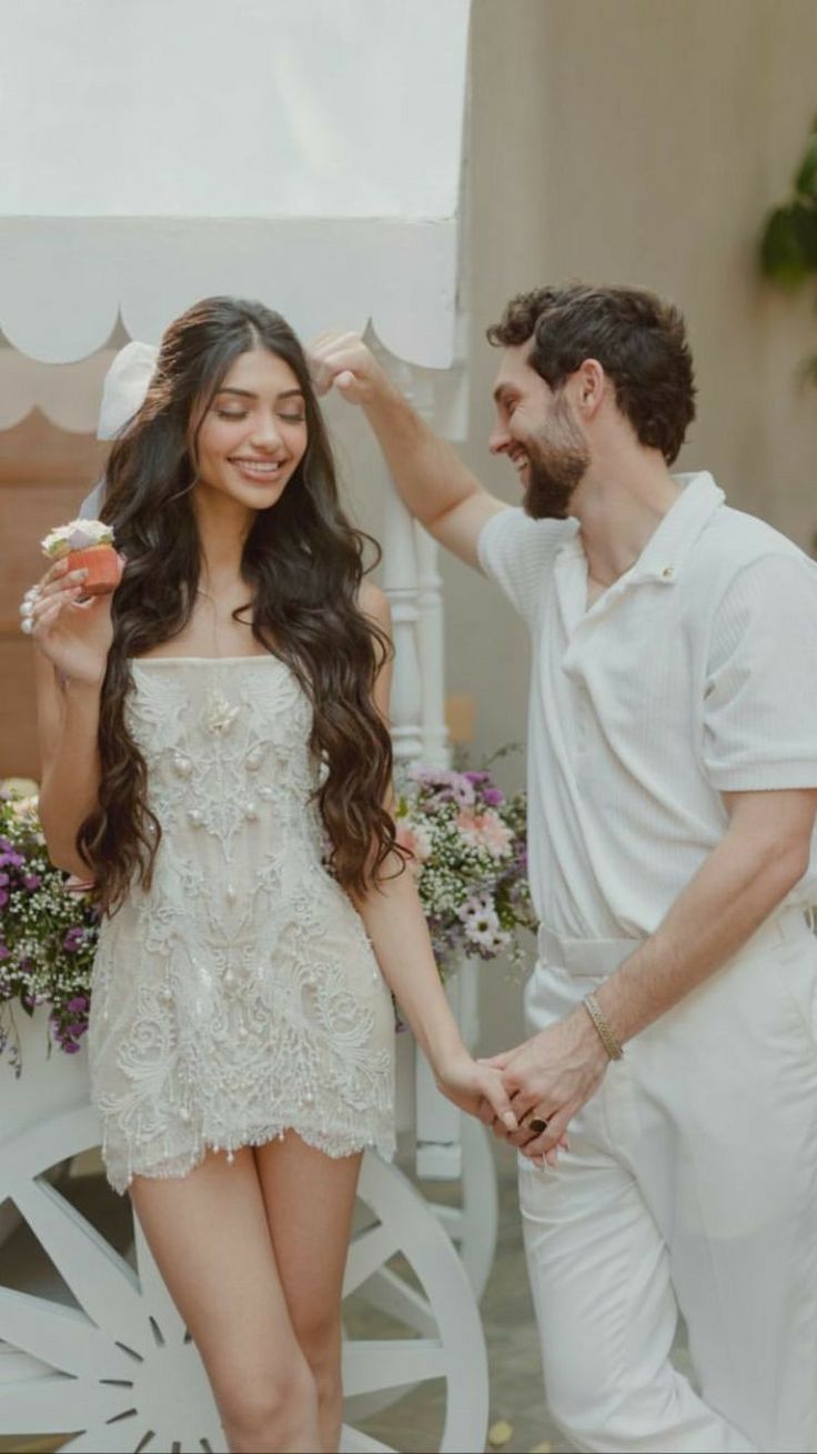 a man standing next to a woman in a white dress and holding hands with her