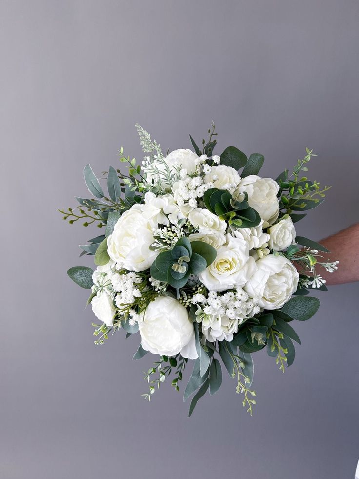 a bouquet of white flowers and greenery is held up by someone's hand